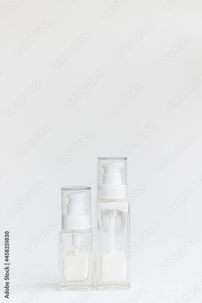 two glass cream bottles with a dispenser lid stand on a white cube. White cream inside transparent containers. Home and professional skin care. two glass cream bottles with a dispenser lid stand on a white cube. White cream inside transparent containers. Home and professional skin care.