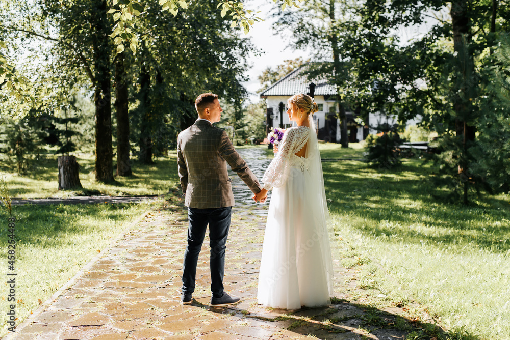 Married young beautiful couple holding hands and looking each other, groom in suit and bride in white dress with bouquet of flowers posing on alley outdoors, back view. Wedding day, marriage concept