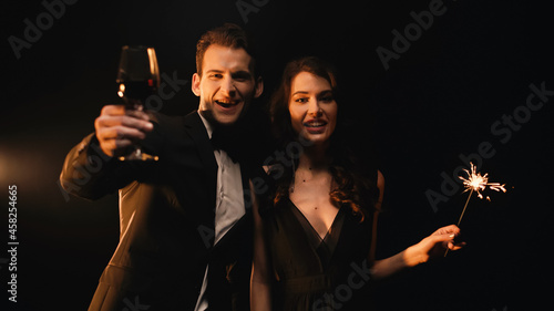 cheerful man holding glass of red wine and woman with sparkler looking at camera on black