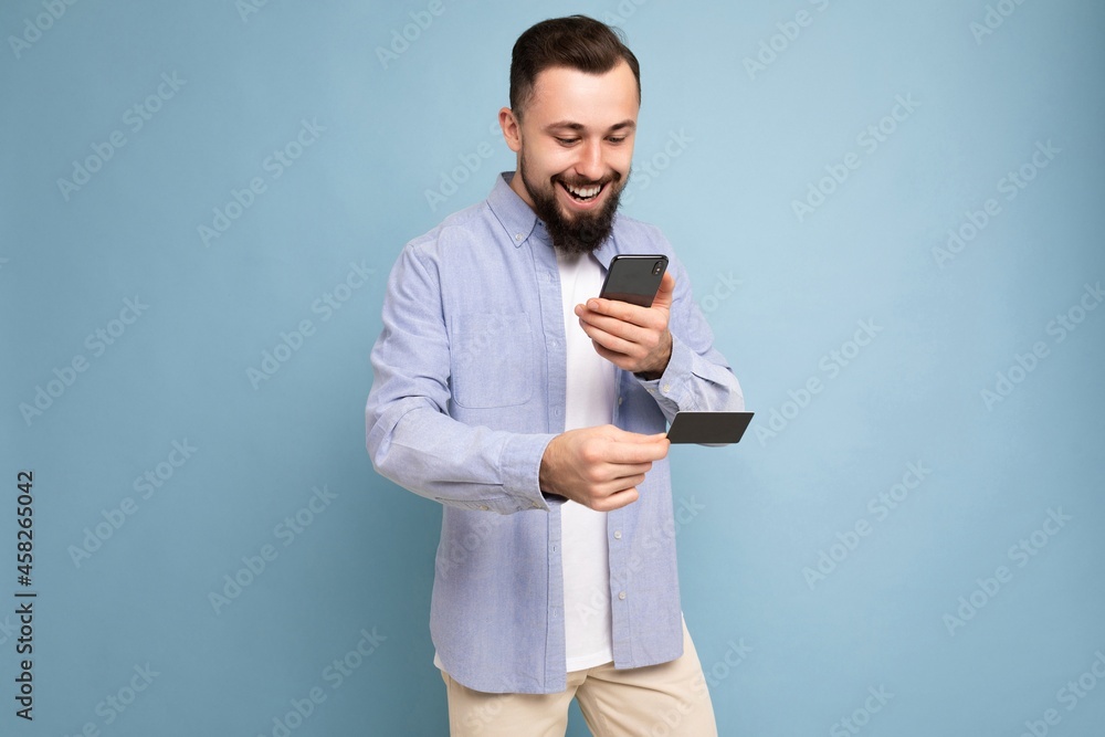 Smiling happy Handsome man wearing everyday clothes isolated on background wall holding and using phone and credit card making payment looking at smartphone screen Smiling happy Handsome man wearing everyday clothes isolated on background wall holding and using phone and credit card making payment looking at smartphone screen