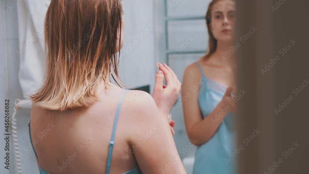 Lovely young elegant caucasian woman in sexy sleepwear looking into window, standing in bathroom and applying facial hydrating creme cosmetics. Beauty treatment. Lovely young elegant caucasian woman in sexy sleepwear looking into window, standing in bathroom and applying facial hydrating creme cosmetics. Beauty treatment.