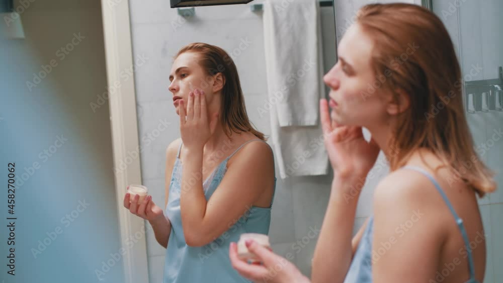 Beautiful woman in bathroom mirror looking on face applying purifying moisturized facial cosmetic cream doing skincare treatment at home.
