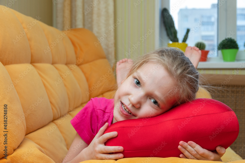 Funny and cute blonde little laughing girl lying on the red pillow on the orange sofa in the children's room. Children's cheerful emotions. Happy child concept.