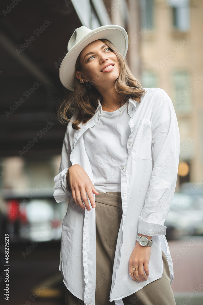 Adorable beautiful young woman expresses happiness, wears fashionable clothes and hat, poses on street, against buildings Adorable beautiful young woman expresses happiness, wears fashionable clothes and hat, poses on street, against buildings