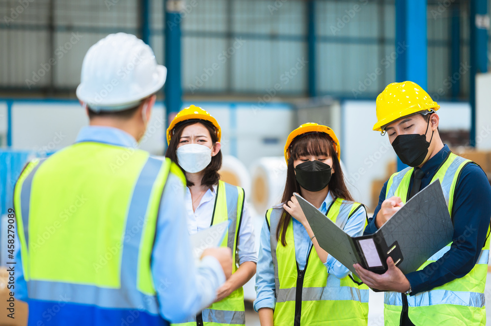The factory employee consists of engineers, foreman, technicians, and related department staff. Wear a mask, hard hat, and vest. meeting before starting work inside the warehouse. Teamwork concept. The factory employee consists of engineers, foreman, technicians, and related department staff. Wear a mask, hard hat, and vest. meeting before starting work inside the warehouse. Teamwork concept.