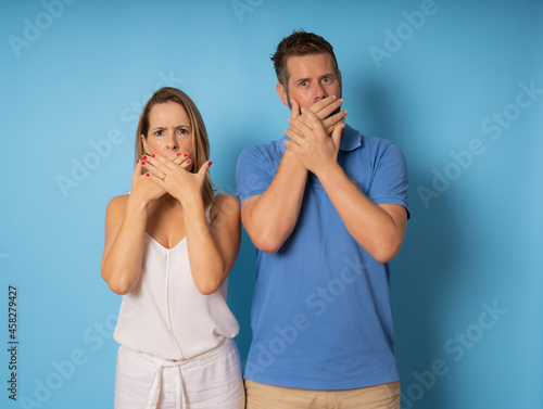 Photo portrait amazed couple keeping secret shut mouth with hands isolated over blue background.