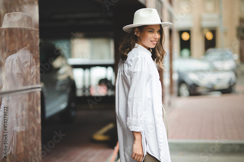 Adorable beautiful young woman expresses happiness, wears fashionable clothes and hat, poses on street, against buildings
