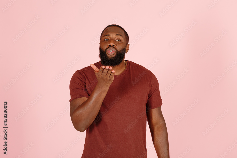 Handsome young African-American man model in casual brown t-shirt sends air kiss standing on light pink background in studio Handsome young African-American man model in casual brown t-shirt sends air kiss standing on light pink background in studio