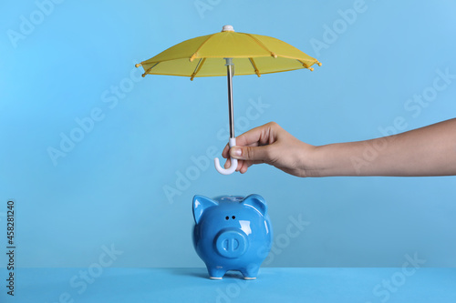 Woman holding small umbrella over piggy bank on light blue background, closeup
