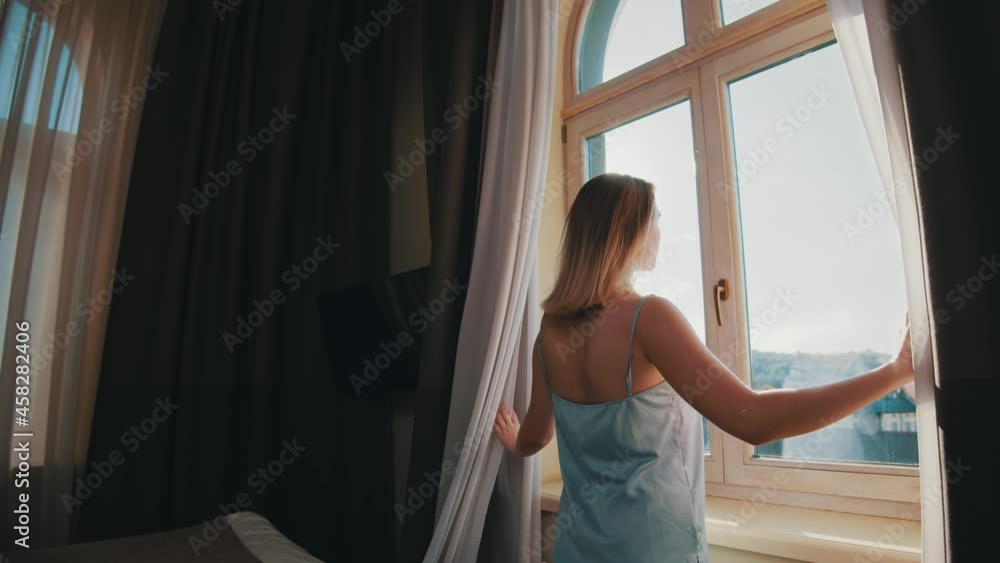 Young happy woman in elegant sleepwear standing by windows to open dark curtains and admire city views. Cityscape in glass window. People in home.