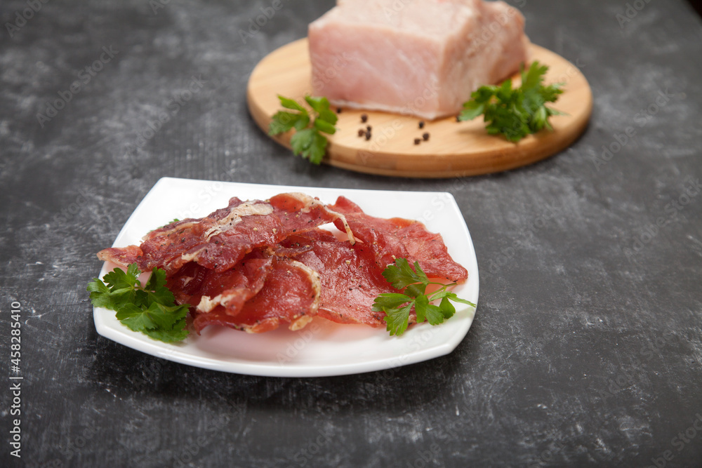 Cured meat on a white plate and raw meat with herbs on a wooden cutting board on a black background.