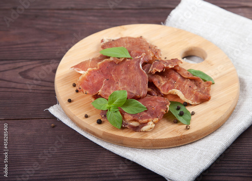 Pieces of jerky with basil leaves lie on a wooden cutting board, under it is a linen napkin on a wooden background.