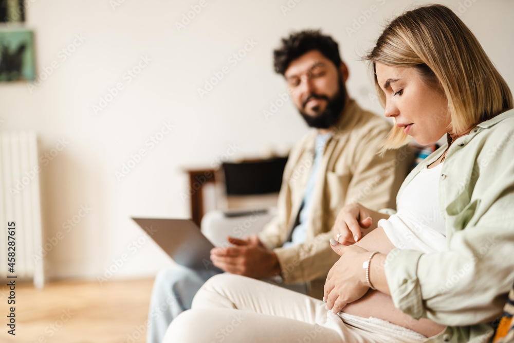 Woman touching her pregnant belly while sitting with her husband on couch Woman touching her pregnant belly while sitting with her husband on couch