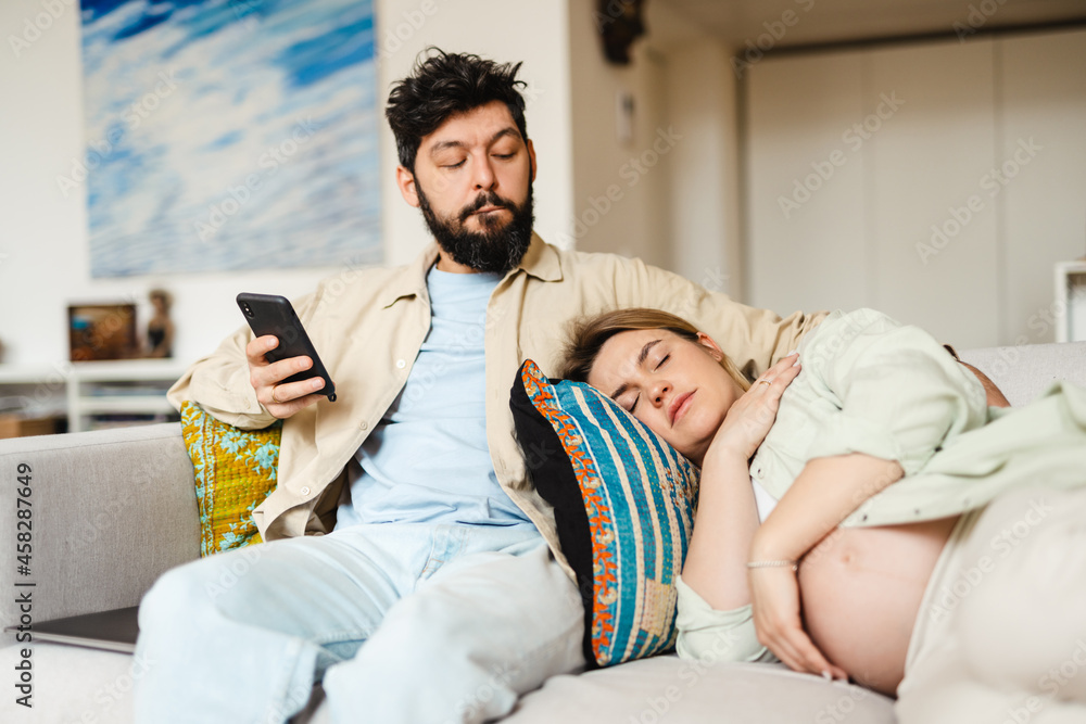 Fototapeta premium Bearded man using cellphone while his pregnant wife sleeping on couch