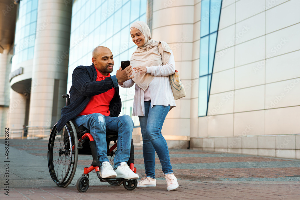 Muslim woman and man in wheelchair using mobile phone together Muslim woman and man in wheelchair using mobile phone together