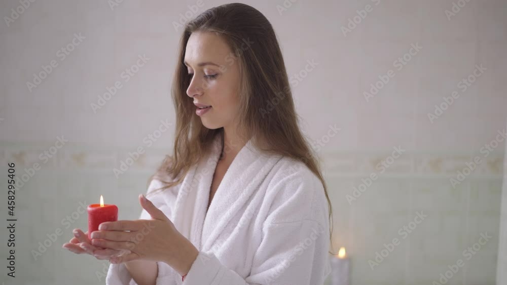 Sensual romantic young woman holding candle standing in bathroom at home dreaming. Portrait of slim beautiful positive Caucasian lady in white bathrobe enjoying leisure indoors. Slow motion Sensual romantic young woman holding candle standing in bathroom at home dreaming. Portrait of slim beautiful positive Caucasian lady in white bathrobe enjoying leisure indoors. Slow motion