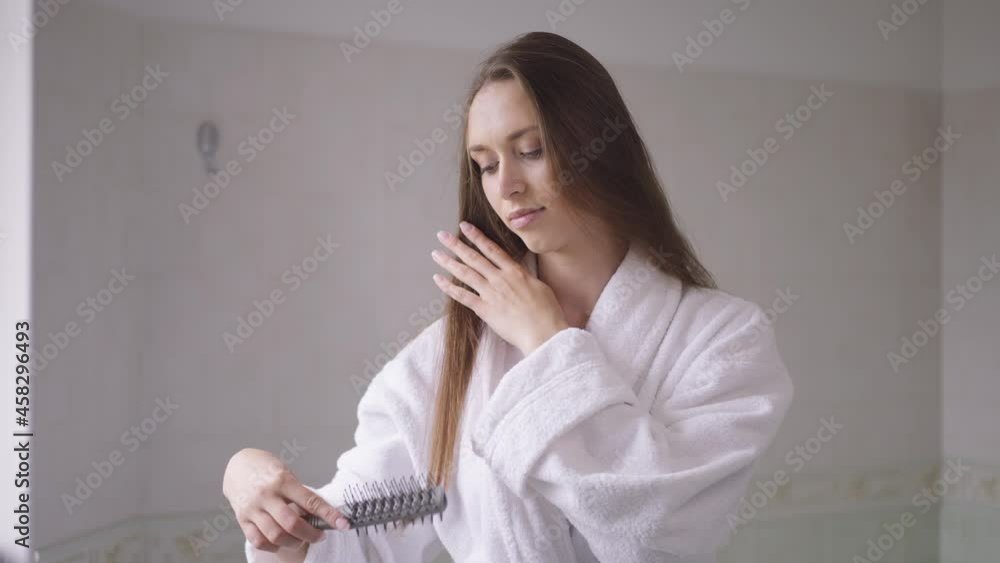 Young smiling Caucasian brunette woman combing hair in slow motion standing at home in bathroom. Portrait of positive confident slim lady enjoying routine in the morning indoors. Lifestyle and beauty Young smiling Caucasian brunette woman combing hair in slow motion standing at home in bathroom. Portrait of positive confident slim lady enjoying routine in the morning indoors. Lifestyle and beauty
