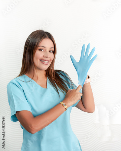 Smiling female doctor in blue uniform  putting on her hand sterile blue latex gloves  in white background