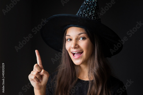 portrait of a smiling girl with her hand pointing up in a witch costume for Halloween close-up on a dark background 