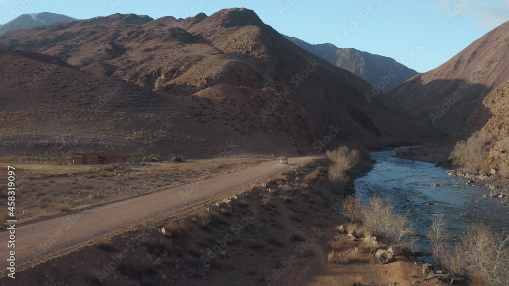 Obraz premium An aerial landscape of a mountain valley with a blue mountain river and a road along which a white car leaves. A picturesque landscape with large mountains against the background of a blue sky.