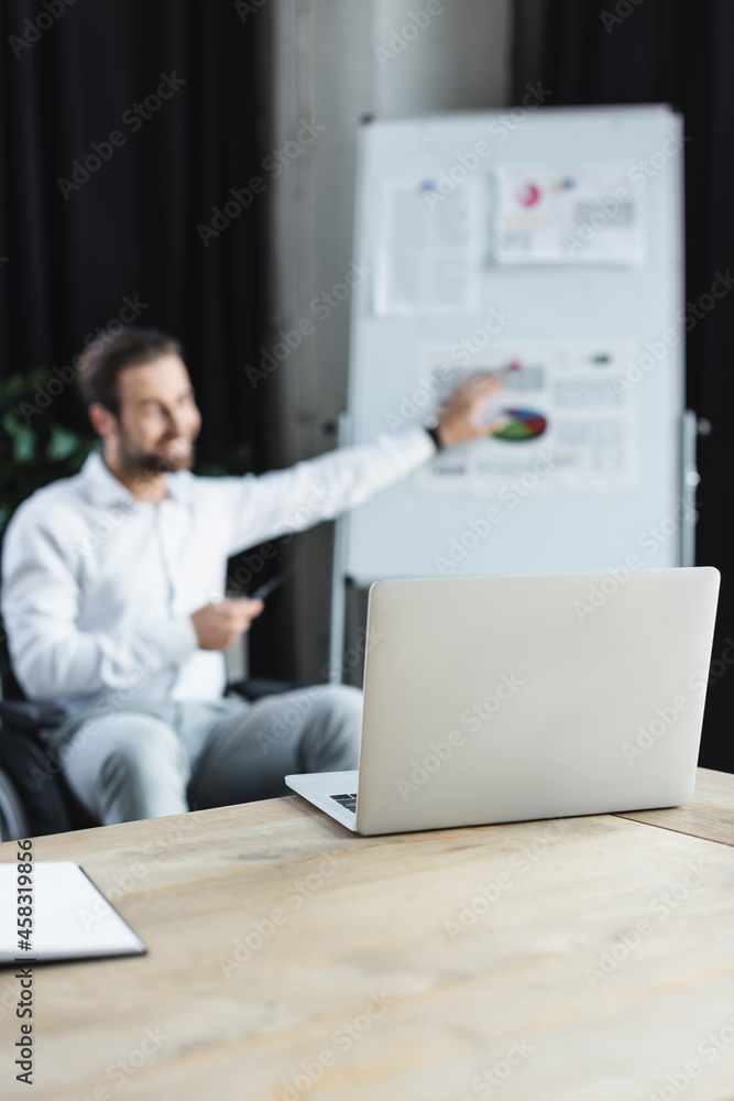 selective focus of laptop near blurred disabled businessman pointing at flip chart during video call selective focus of laptop near blurred disabled businessman pointing at flip chart during video call