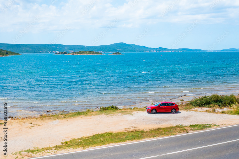 The red car is parked by the sea with a beautiful mountain view