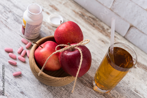 Three fresh red apples in a wooden bowl, glass of juice, scattered pills and jar of pills. Choice of healthy life