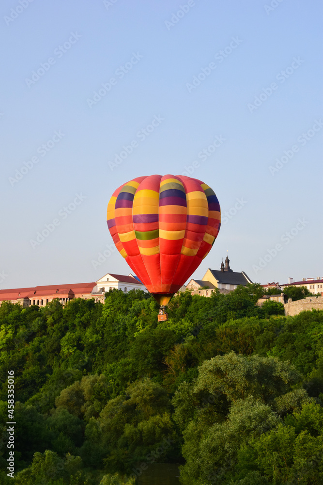 Fototapeta premium A large multi-colored balloon flies very low over the river and between dense trees. In the background, the buildings of the ancient city