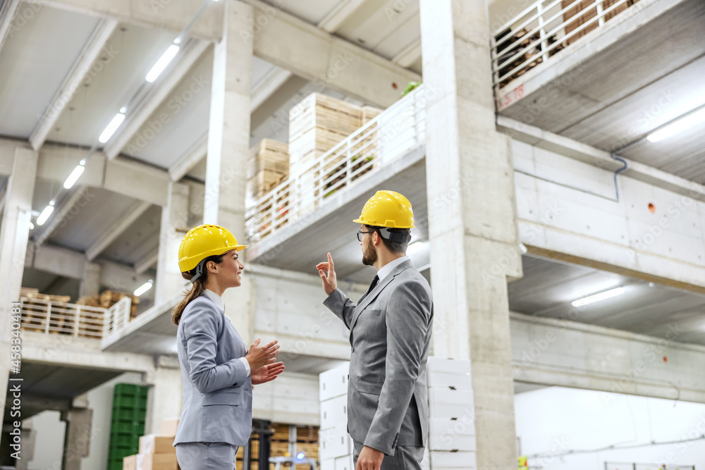 Warehouse and management. A man and a woman wear a business suit and helmet in a warehouse and talking about work. An adult woman watches a man as he explains and points to something on the side Warehouse and management. A man and a woman wear a business suit and helmet in a warehouse and talking about work. An adult woman watches a man as he explains and points to something on the side