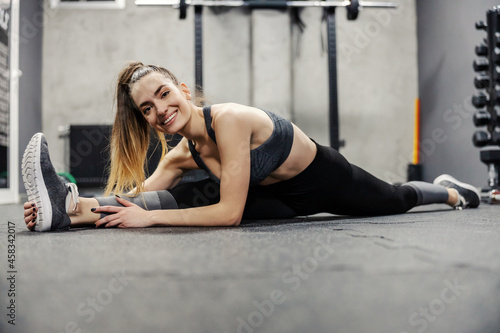 Muscle flexibility and hip mobility. Fit woman in sportswear and good body shape stretches legs muscles on the floor of an isolated gym. Enjoyment and smile in training, ponytail. Looking at camera
