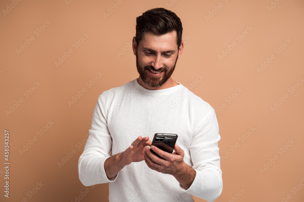 Assertive bearded man attentively looking at display of smartphone he holding, surfing internet, doing shopping online. Indoor studio shot isolated on beige background Assertive bearded man attentively looking at display of smartphone he holding, surfing internet, doing shopping online. Indoor studio shot isolated on beige background
