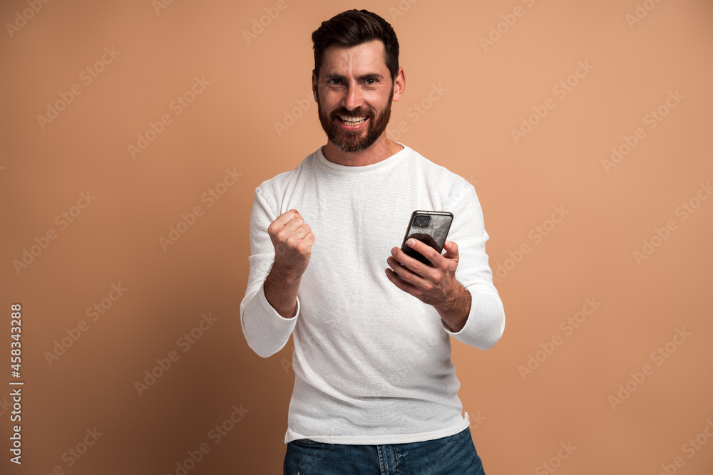 Happy satisfied man with beard holding smartphone and smiling making yes gesture, celebrating online lottery or giveaway victory. Indoor studio shot isolated on beige background Happy satisfied man with beard holding smartphone and smiling making yes gesture, celebrating online lottery or giveaway victory. Indoor studio shot isolated on beige background