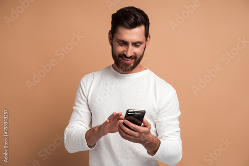 Assertive bearded man attentively looking at display of smartphone he holding, surfing internet, doing shopping online. Indoor studio shot isolated on beige background © speed300