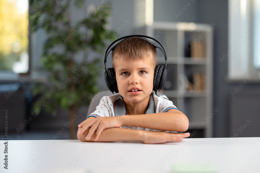 Portrait of caucasian schoolboy sitting at table in wireless headphones. Cute boy having online lesson while staying at home. Distance learning concept. Portrait of caucasian schoolboy sitting at table in wireless headphones. Cute boy having online lesson while staying at home. Distance learning concept.