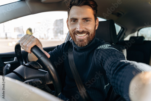 Great selfie. Handsome bearded man holding camera and making selfie while sitting in the car at the driver seat and looking at the camera with pleasure smile