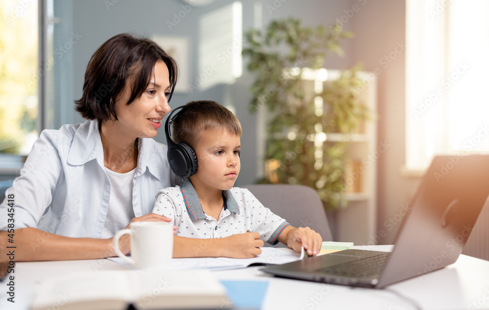 Little schoolboy in headphones sitting at desk and looking at computer screen during online lesson. Caring mother sitting near and helping her son studying at home. Little schoolboy in headphones sitting at desk and looking at computer screen during online lesson. Caring mother sitting near and helping her son studying at home.