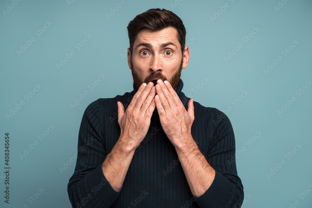 Shocked man closing mouth with hands, confused and depressed with information, looking with astonishment and fear, privacy. Indoor studio shot isolated on blue background Shocked man closing mouth with hands, confused and depressed with information, looking with astonishment and fear, privacy. Indoor studio shot isolated on blue background