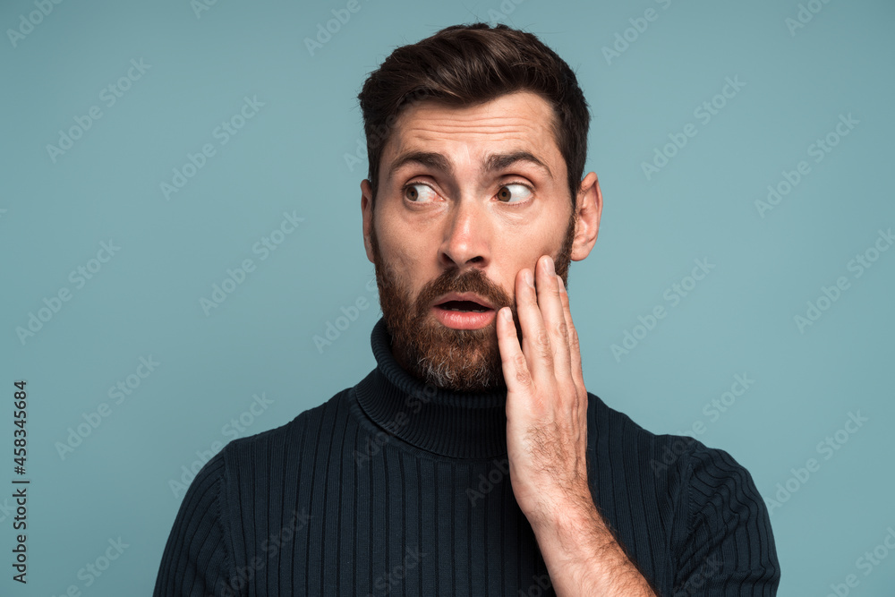 Excited shocked man holding hands on face and looking away with big eyes and open mouth, shocked bemused with news, win at lottery. Indoor studio shot isolated on blue background