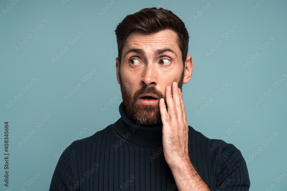 Excited shocked man holding hands on face and looking away with big eyes and open mouth, shocked bemused with news, win at lottery. Indoor studio shot isolated on blue background Excited shocked man holding hands on face and looking away with big eyes and open mouth, shocked bemused with news, win at lottery. Indoor studio shot isolated on blue background