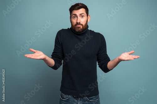 I don't know. Portrait of confused handsome bearded young man standing with raised arms and looking at camera with answer. Indoor studio shot, isolated on blue background