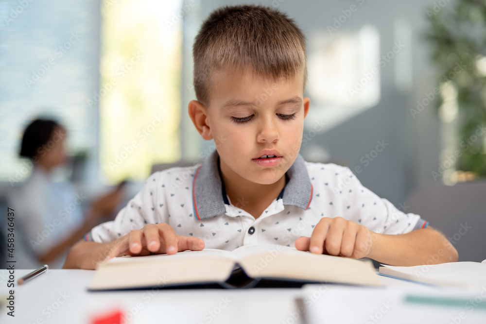 Focused little boy sitting at desk and reading book. Caucasian schoolboy spending free time for studying at home. Concept of domestic learning. Focused little boy sitting at desk and reading book. Caucasian schoolboy spending free time for studying at home. Concept of domestic learning.