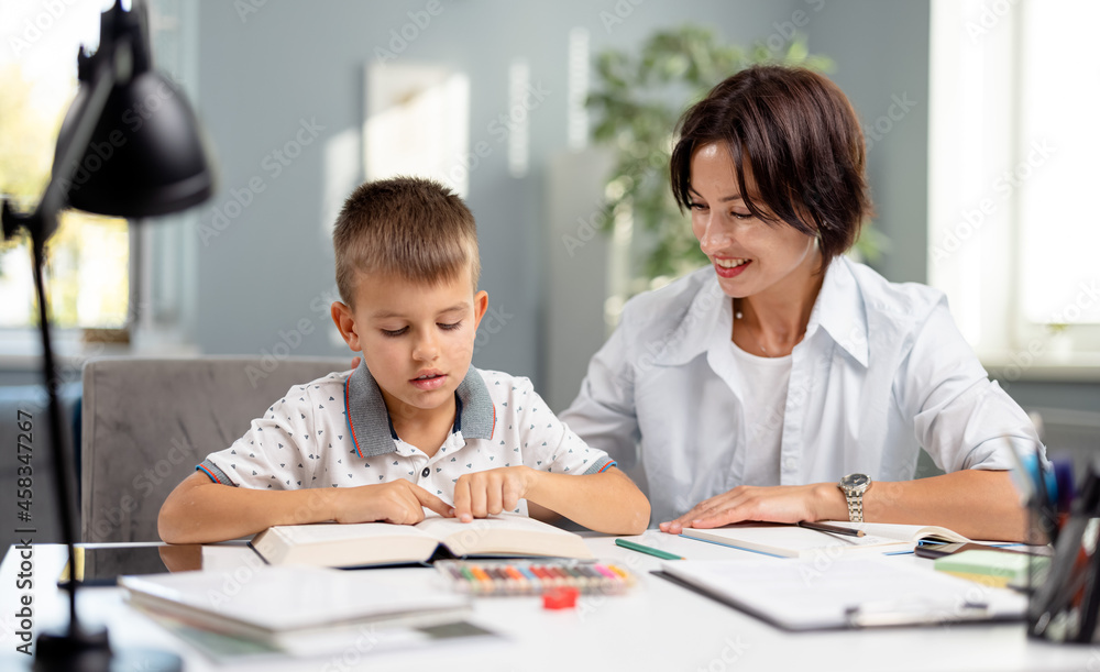 Caucasian schoolboy sitting with mother at desk and reading book. Caring woman helping son with homework. Educational moments during parenting. Caucasian schoolboy sitting with mother at desk and reading book. Caring woman helping son with homework. Educational moments during parenting.