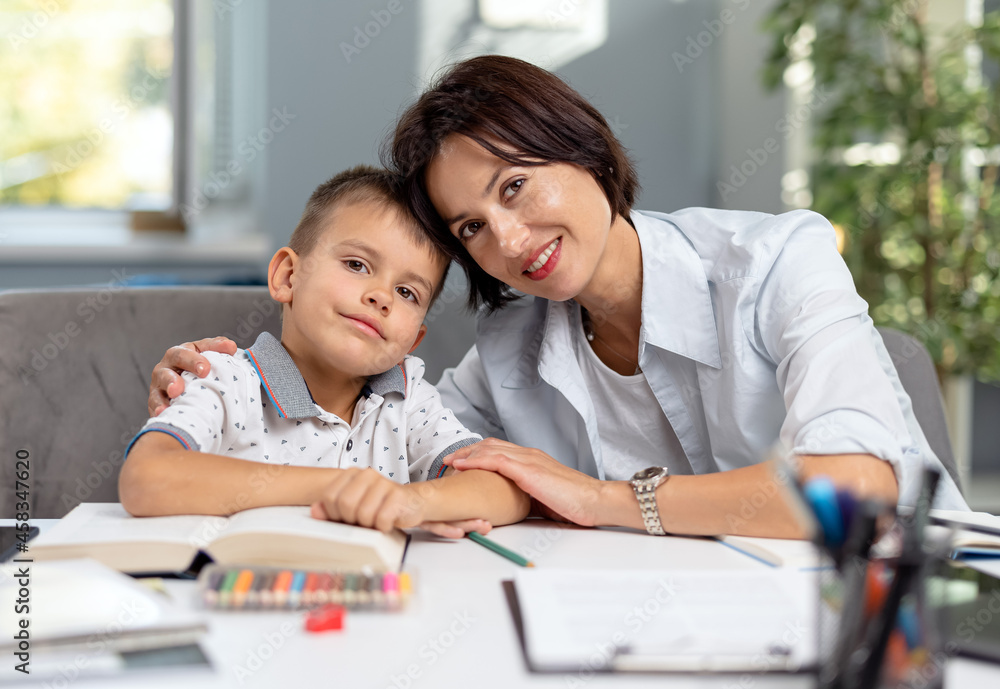 Happy caucasian mother embracing her lovely son while sitting together at desk with books and notes. Domestic studying and family time concept. Happy caucasian mother embracing her lovely son while sitting together at desk with books and notes. Domestic studying and family time concept.