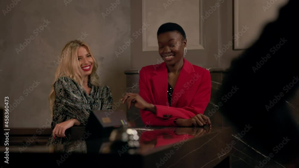 Two Cheerful Girls In Evening Dresses Talk At Reception At Hotel. Two Cheerful Girls In Evening Dresses Talk At Reception At Hotel.
