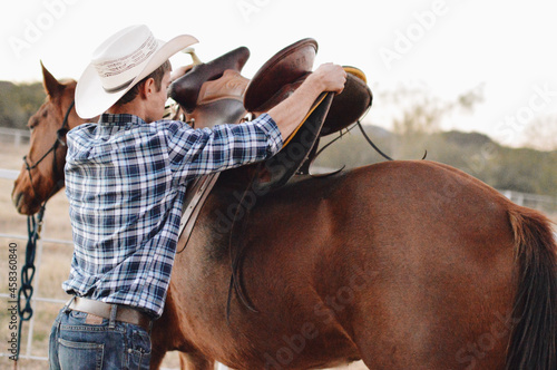 Fototapeta Male farmer in a cowboy hat attaching a saddle onto a brown horse