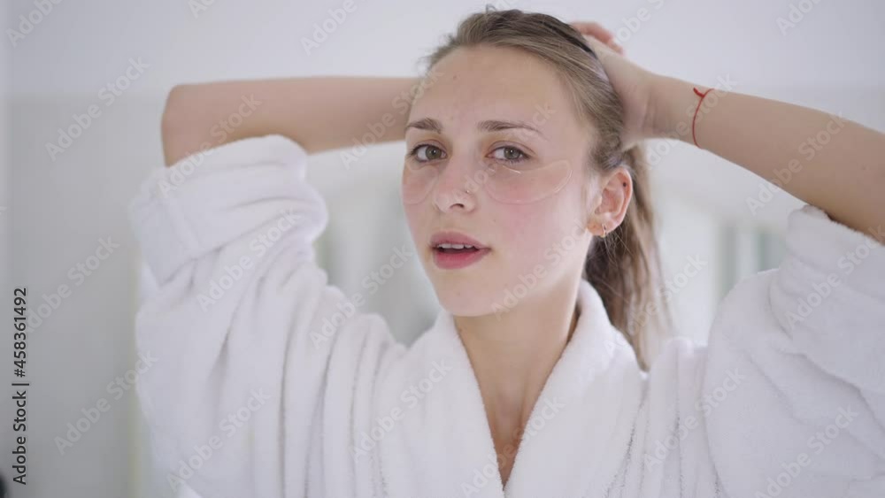 Gorgeous slim young woman with eye patches on face making ponytail looking at camera. Portrait of beautiful slim Caucasian millennial lady taking care of facial skin at home. Morning beauty routine Gorgeous slim young woman with eye patches on face making ponytail looking at camera. Portrait of beautiful slim Caucasian millennial lady taking care of facial skin at home. Morning beauty routine
