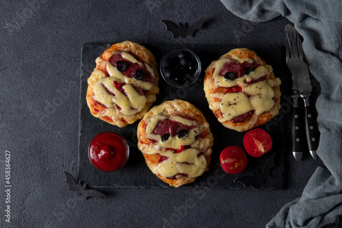 Funny mini pizzas mummies with sausage and cheese for Halloween party, served with black olives, ketchup and tomatoes on dark background