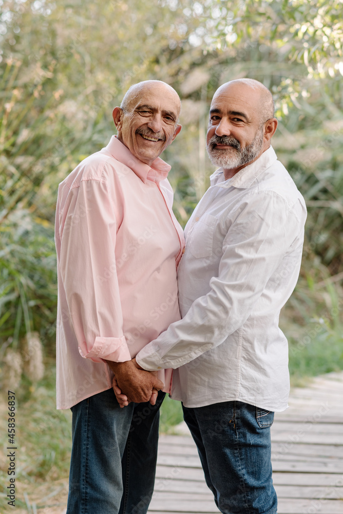 Happy middle-aged gay couple holding hands and looking at the camera while enjoying a walk in the park together. LGBT couple concept.