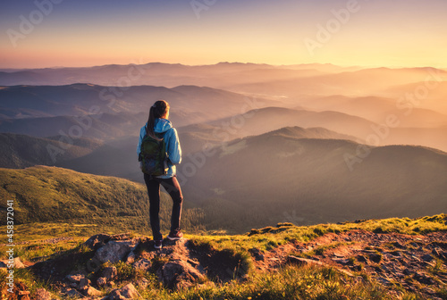 Girl with backpack on mountain peak with green grass looking in beautiful mountain valley in fog at sunset in autumn. Landscape with sporty young woman, foggy hills, orange sky in fall. Hiking. Nature
