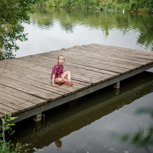 A blonde girl sits on a wooden pier by the river.